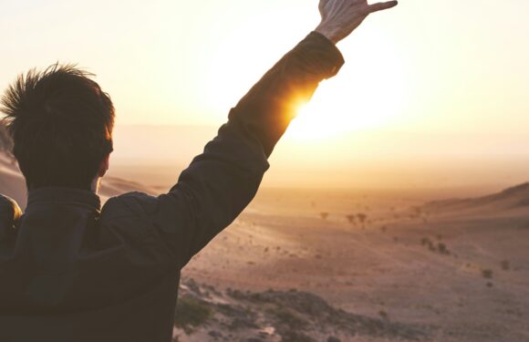 A person in a dark jacket raises one arm towards the sunrise over a desert landscape with sand dunes and sparse vegetation.