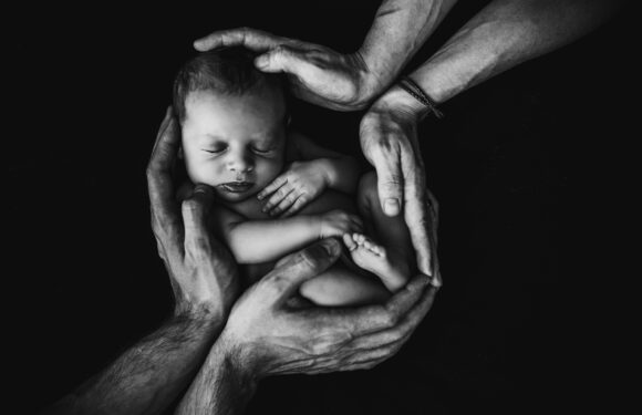 A newborn baby peacefully sleeping in a tiny curled-up position, cradled and gently held by multiple adult hands against a black background.