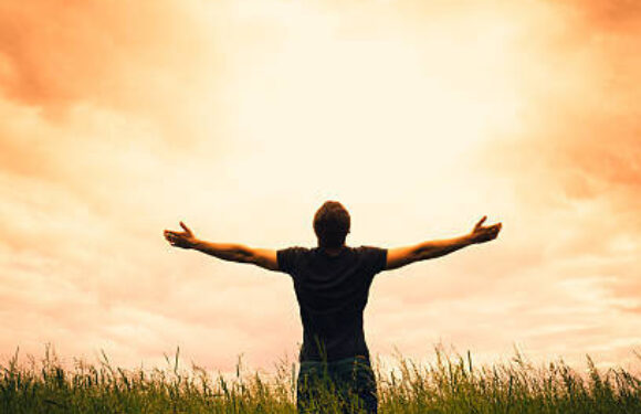 Person standing in tall grass with arms outstretched under a glowing orange sunset sky.