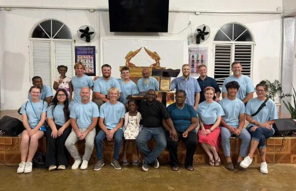Group of people, mostly in light blue shirts, posing on a stage in a room with white walls, arched windows, and fans, with a framed document and decorative figure in the background.