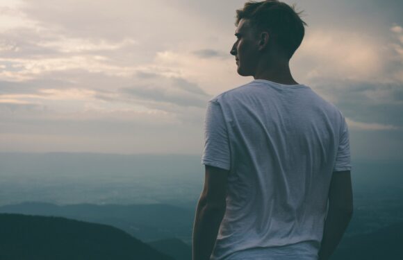 A person in a light-colored t-shirt standing outdoors during sunset, looking towards a vast landscape of rolling hills and sky with scattered clouds.