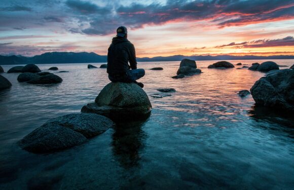 Person sitting on stacked rocks by a calm sea at sunset, wearing a cap and dark jacket as waves reflect colorful sky.