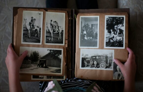 Hands holding an old photo album with several black-and-white pictures of people, horses, a bicycle, and an early car