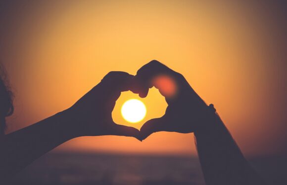 Silhouetted hands forming a heart shape with a glowing sun between fingers at sunset, warm orange and purple sky background