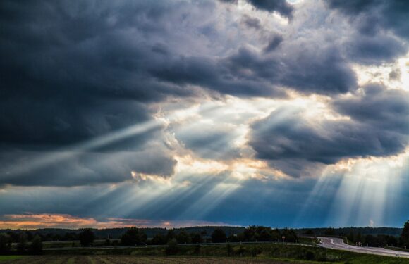 Dark, cloudy skies with sunlight rays breaking through, over a rural landscape with a winding road and distant trees.