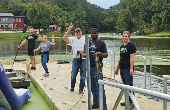 A group of five people on a dock by the water, some smiling, one paddling a kayak, with trees and colorful buildings in the background.