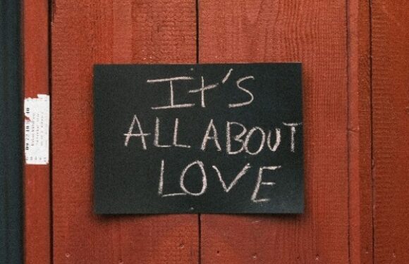 Handwritten chalk message on a blackboard sign reading “IT’S ALL ABOUT LOVE” on a reddish wooden door.