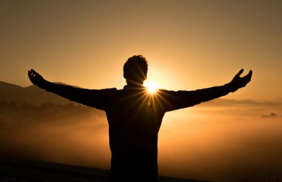 Silhouette of a person with arms outstretched facing a sunrise over misty mountains and a golden sky