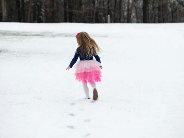 A young girl with long hair in a pink bow, wearing a dark top, pink tutu, and white tights, walks away across snow with a forest in the background.