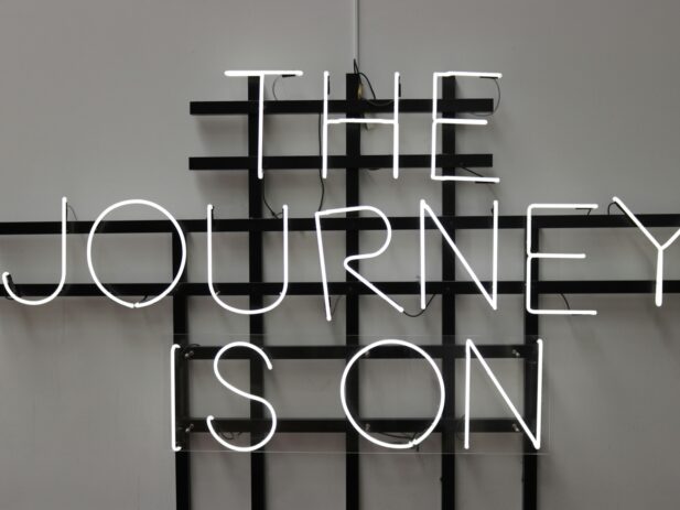 A neon sign on a gray wall displaying the words "THE JOURNEY IS ON" with a black grid structure behind the sign.