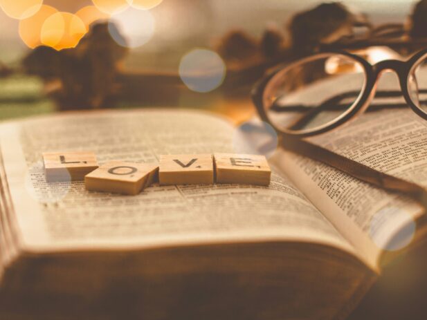 An open Bible with wooden tiles spelling "LOVE" placed on it, with a pair of glasses resting on the right page, warm lighting, and blurred background.