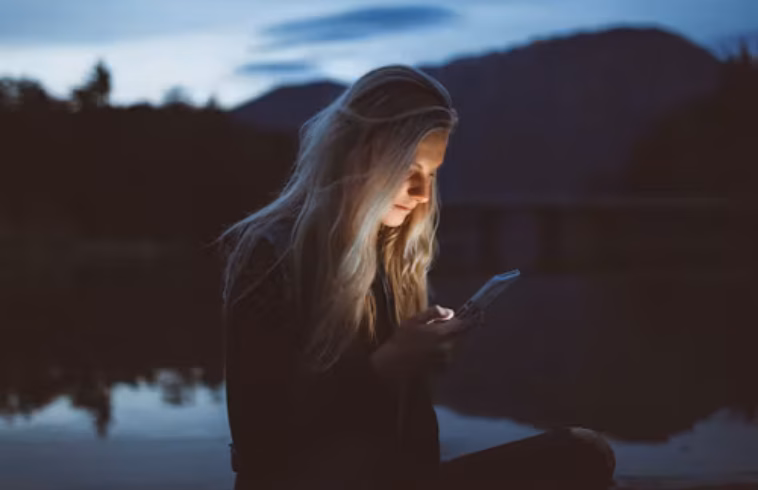 Woman with long hair wearing a cap, sitting by a calm lake at dusk and looking at a smartphone, with mountains in the background.
