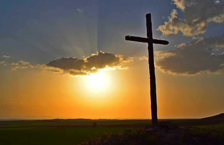 Dark cross silhouette on a hillside at sunset, glowing clouds and golden sky with sun rays behind it.