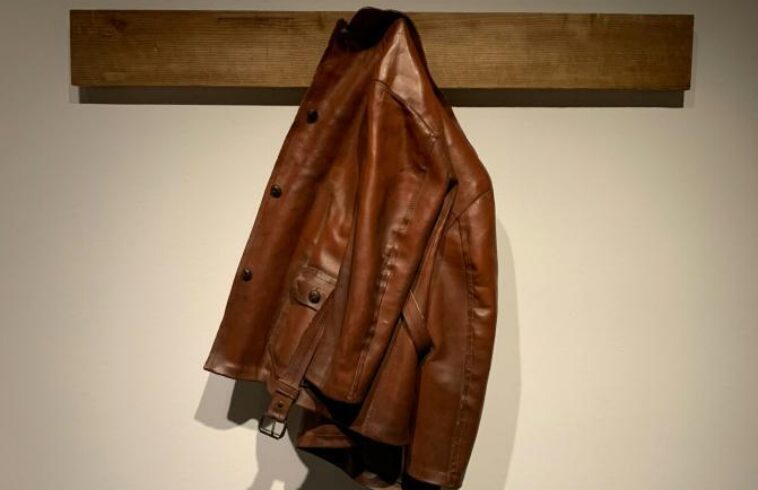 Brown leather jacket hanging on a wall mount beam against a plain light background.