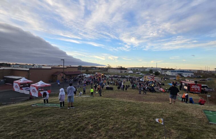 Crowd gathers on a grassy field at an outdoor fun event near buildings, with food booths and trucks under a sunset sky.
