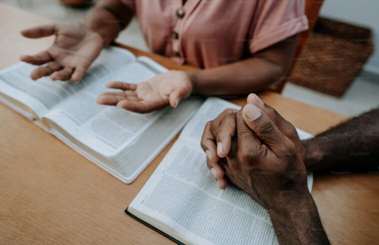 Two people are engaged in a conversation over open books on a wooden table; one person's hands are clasped, while the other's hands are gesturing as they speak.