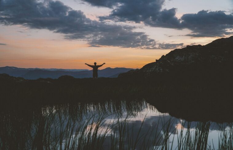 A person stands with arms outstretched on a hill at sunset, with a mountain range behind them, a small body of water reflecting the sky and grass in the foreground.