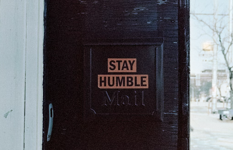 A dark door with a sign that reads "STAY HUMBLE" in uppercase letters, located in a building with light-colored siding; a street scene with trees and buildings is visible in the background.