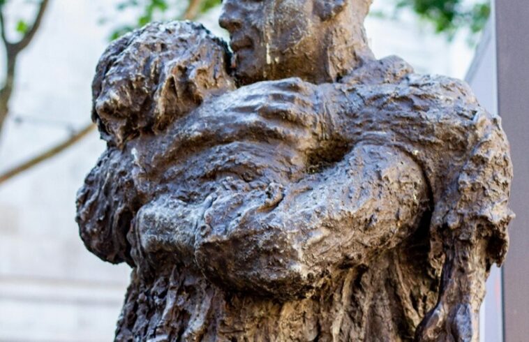 Bronze statue of a kneeling person holding a child figure, outdoors with trees blurred in the background.