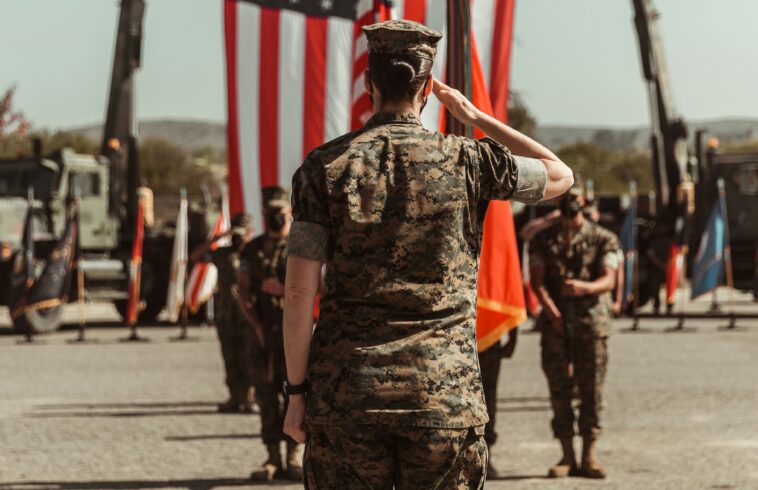Soldier in camouflage standing in front of a large American flag during an outdoor ceremony with uniformed troops in the background.