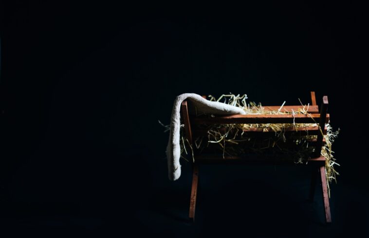 A small wooden bench with hay scattered on top, a white cloth draped over one side, set against a dark background with minimal lighting.