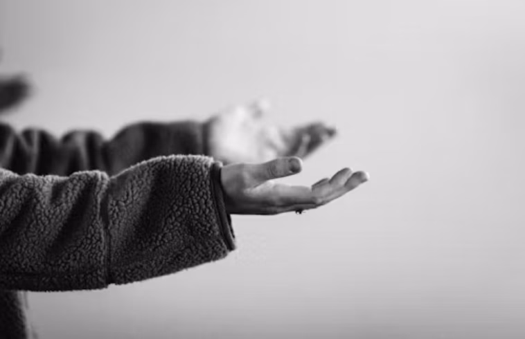 Close-up of a person’s hands in a dark sleeve, palm open and reaching, with a blurred background and light gray tones.