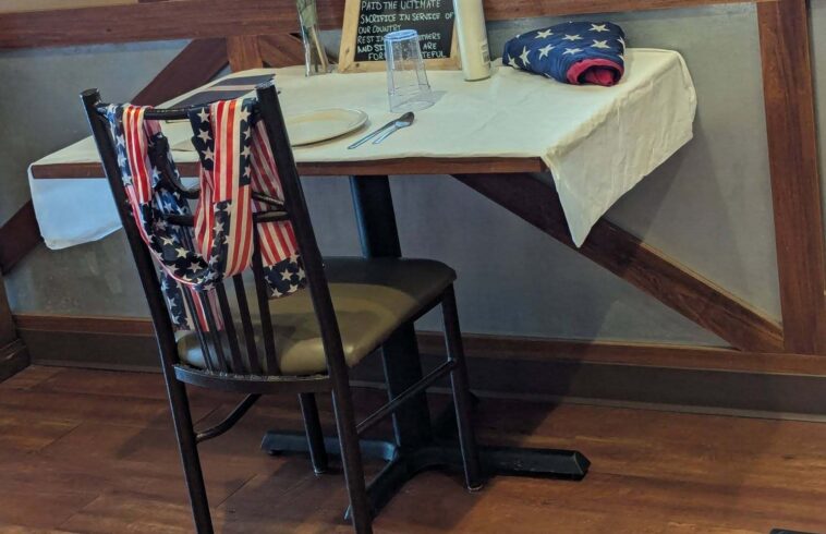 Dining setup with small table covered in white cloth, American flags draped over chairs, bunting and a “fallen comrade” sign by a window.