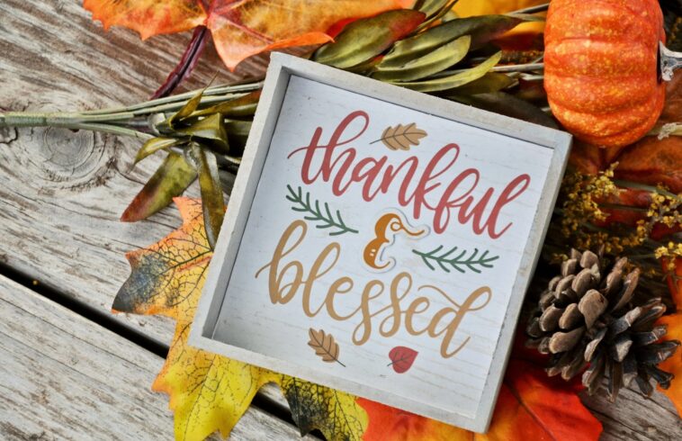 A decorative fall-themed sign on a wooden surface surrounded by colorful autumn leaves, small pumpkins, and pinecones with a cheerful "thankful & blessed" message.