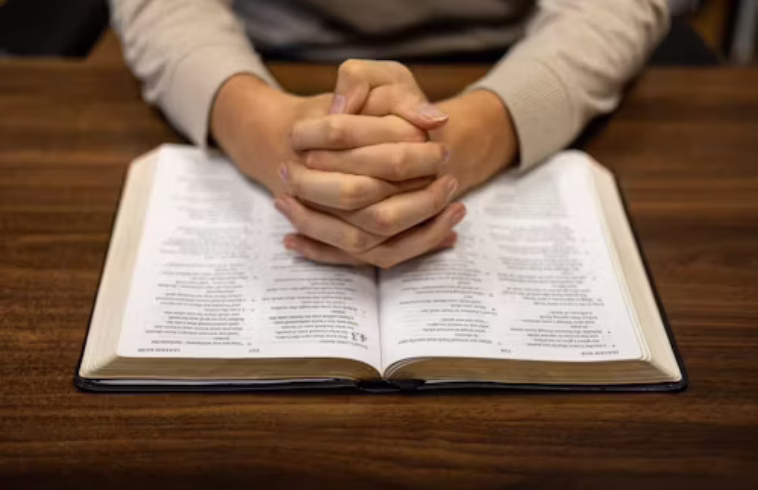 Person with hands clasped over an open Bible on a wooden table, wearing a beige sweater.