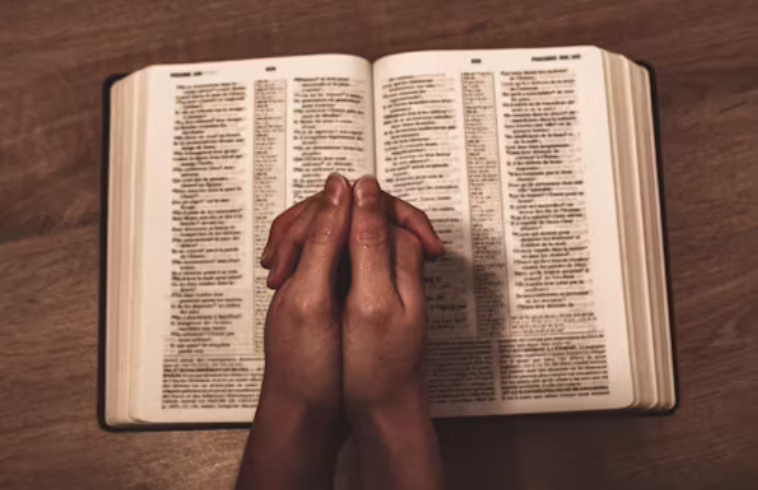 A person praying with clasped hands over an open Bible on a wooden surface.