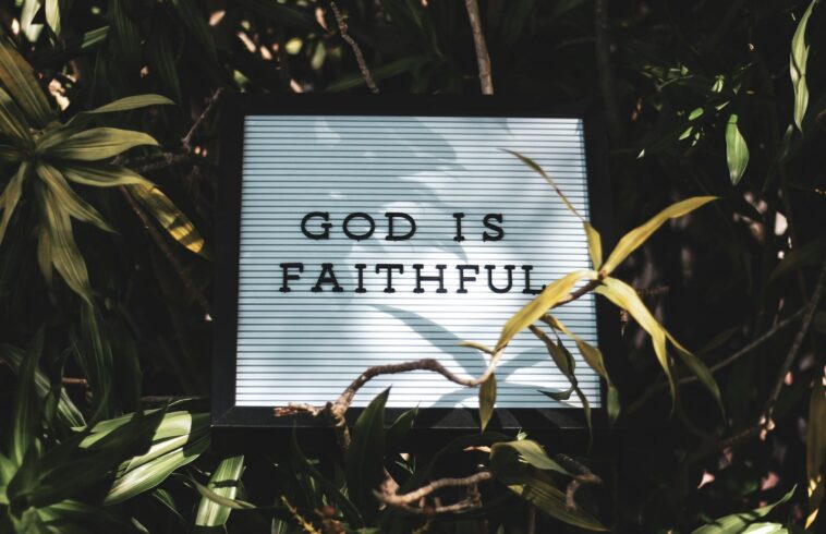 Backlit sign reading “GOD IS FAITHFUL” framed by tropical leaves and branches in warm sunlight.