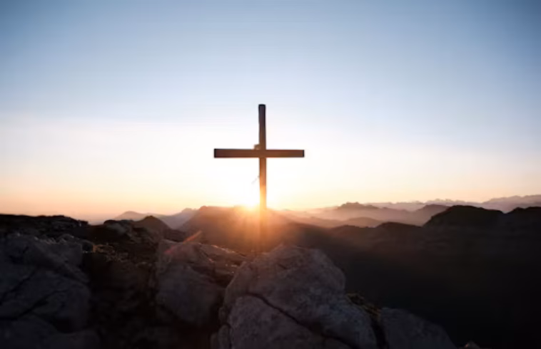 Sunrise over rugged mountain peaks with a wooden cross silhouetted on a rocky summit, glowing sky behind.
