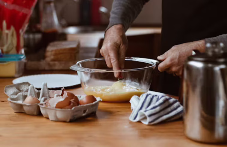 Person cracking eggs into a glass bowl on a wooden counter, with eggs in a carton nearby, a towel, and a stainless steel container nearby.