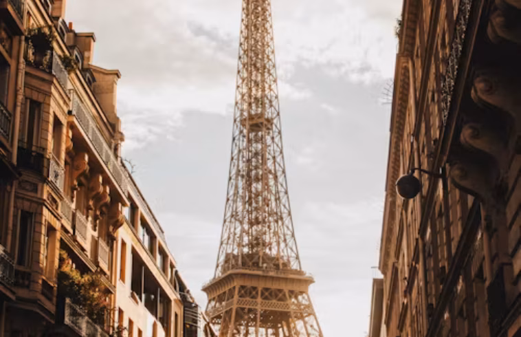 Eiffel Tower rising between tall Parisian buildings at sunset, viewed from a narrow street with warm light and cloudy sky