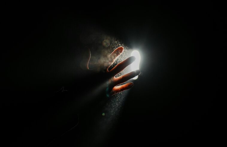 Hand reaching into glowing mist, fingertips lit with sparks and water droplets against a dark background, dramatic lighting.