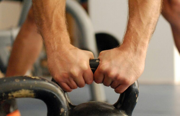 Two bare arms gripping a black kettlebell handle inside a gym, with background people and equipment blurred