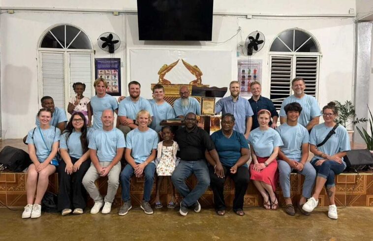 Group of people, mostly in light blue shirts, posing on a stage in a room with white walls, arched windows, and fans, with a framed document and decorative figure in the background.