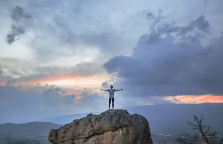 Person standing on a rocky hilltop with arms outstretched at sunset, dramatic clouds over distant mountains.