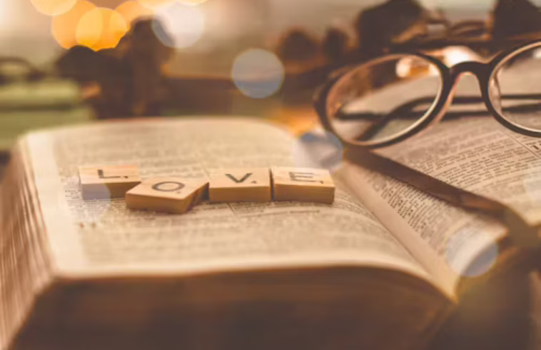 A Bible open with wooden blocks spelling "LOVE" on the pages, a pair of glasses resting nearby, with a warm, blurred background.