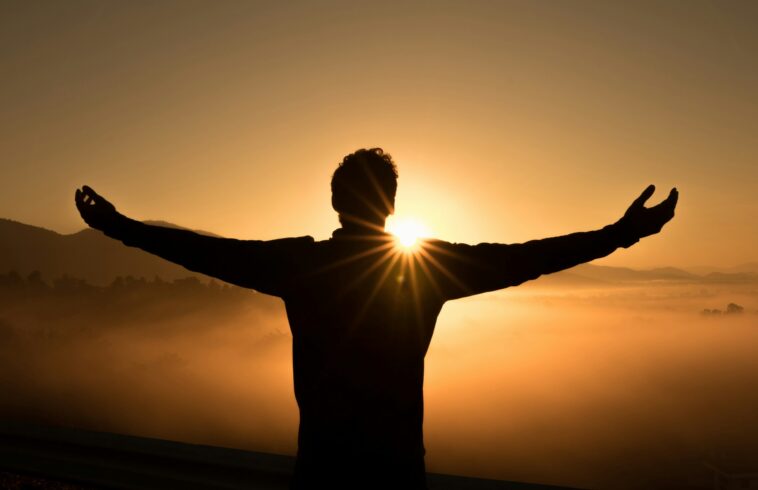 Silhouette of a person with arms outstretched facing a sunrise over misty mountains and a golden sky