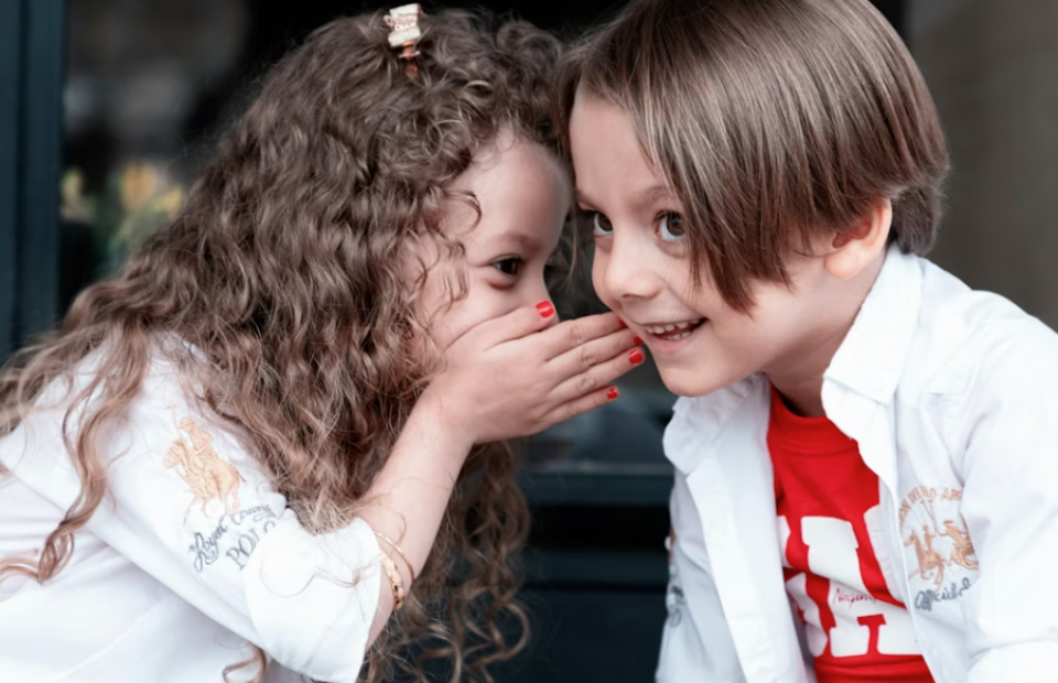 Two children, a girl with curly hair and a boy with straight hair, sharing a secret outside. Both are smiling, and the girl covers her mouth with her hand.