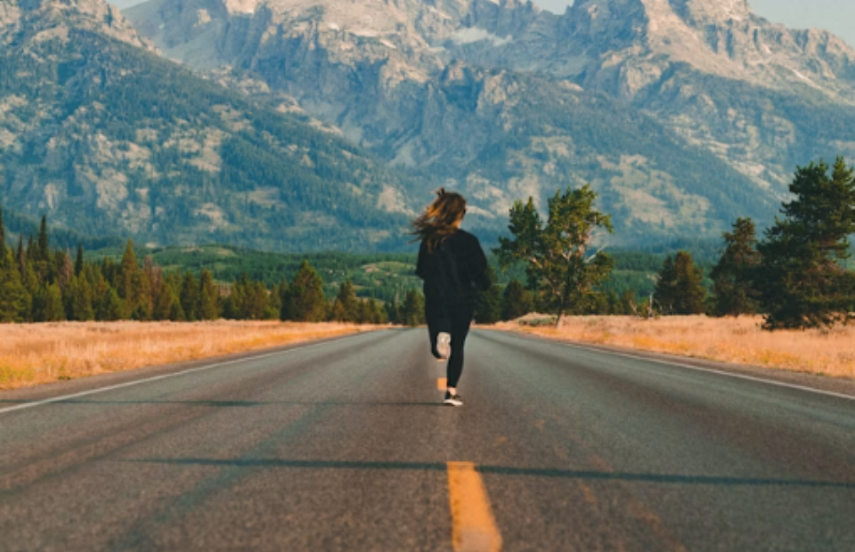 A person walking on an empty road surrounded by fields with mountains in the background during daytime.
