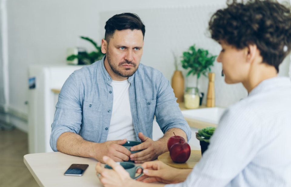 A man with a serious expression wearing a light blue shirt sits across from a woman with curly hair, holding a mug, in a bright, modern kitchen.