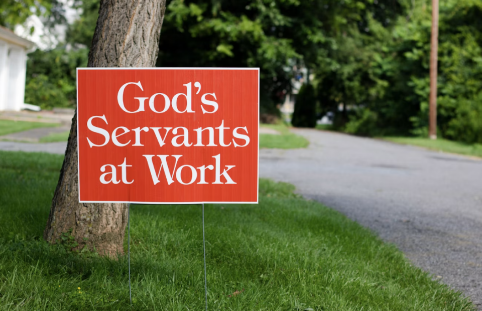 Red sign on a tree trunk in a green yard reading “God’s Servants at Work,” with a paved road and trees in the background