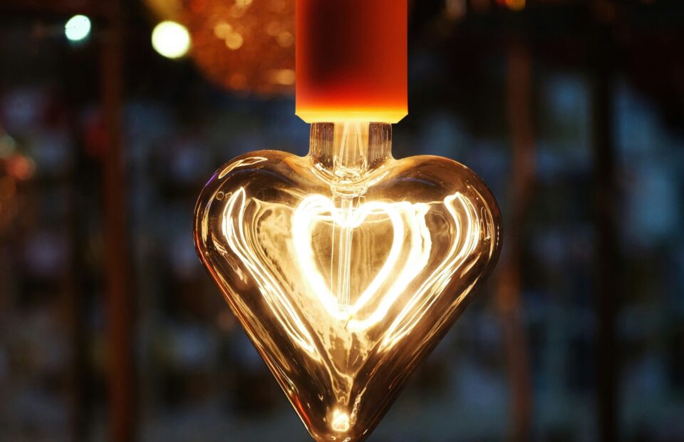 A glowing light bulb shaped like a heart, with an inner heart design illuminated by the filament, hanging outdoors with a blurred background of lights.