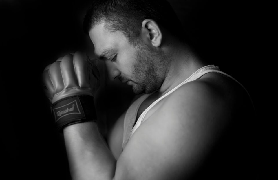 Black-and-white portrait of a person with a boxing glove, head bowed and eyes closed, softly lit against a dark background.