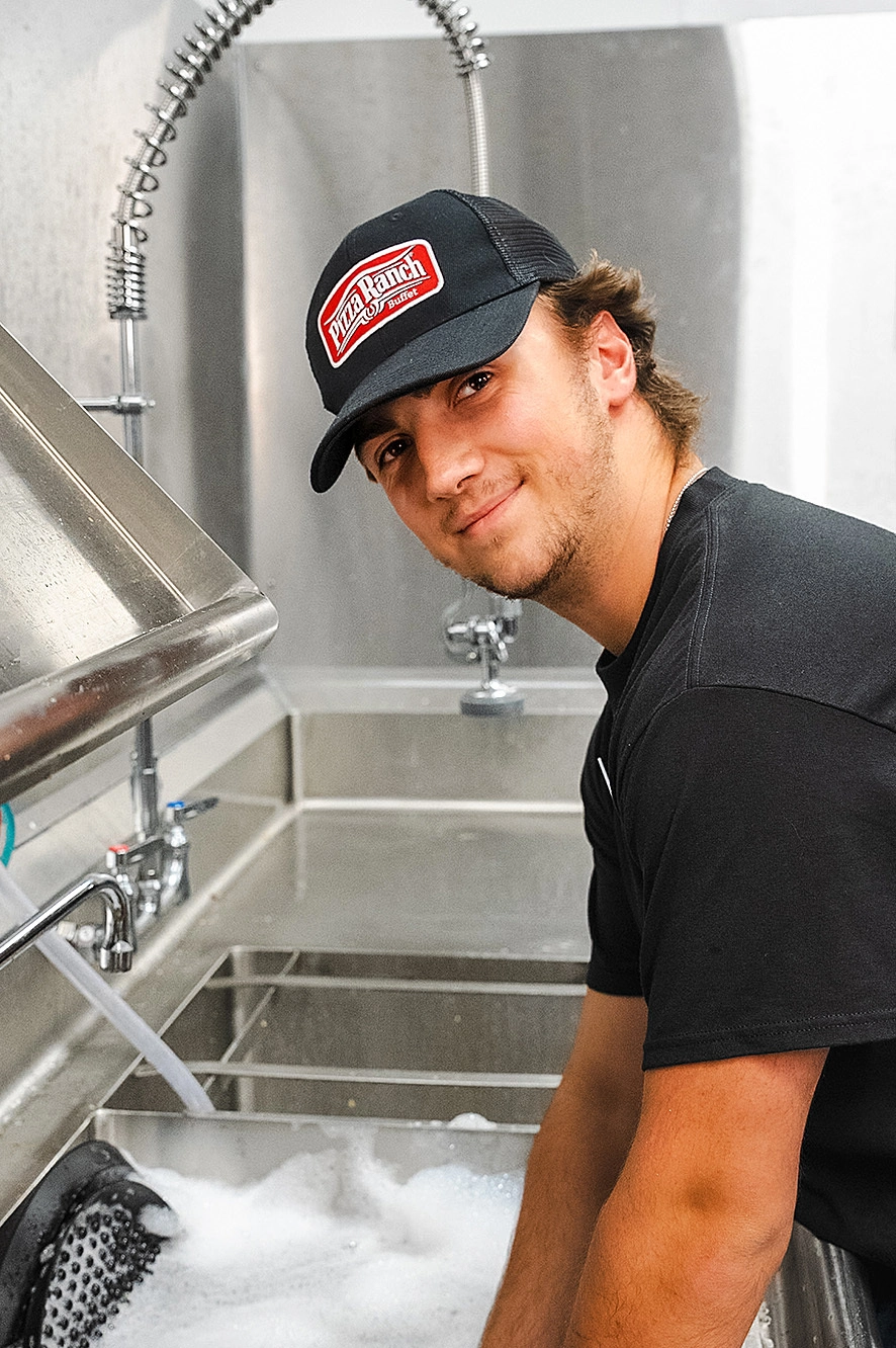 Young man wearing a black Pizza Ranch hat washing dishes in a commercial sink with soapy water. Gray industrial kitchen background.