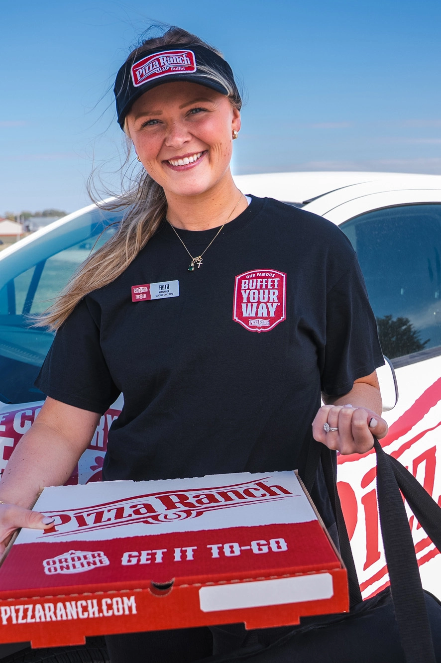 A smiling woman wearing a black Pizza Ranch uniform and visor holds a pizza box, standing outdoors near a white vehicle with a clear blue sky in the background.