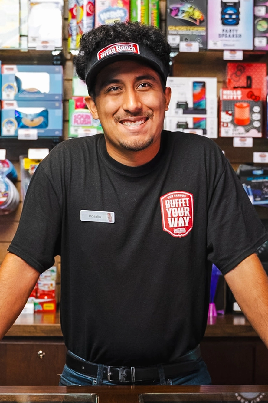 A smiling man wearing a black Pizza Ranch uniform and a visor stands behind a counter in a store. The background features shelves with colorful products.