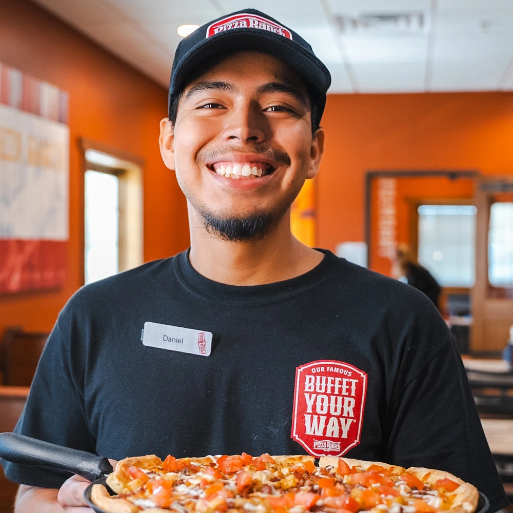 Smiling employee in a black Pizza Ranch shirt and cap, holding a freshly delivered pizza, inside a brightly lit restaurant with orange walls.